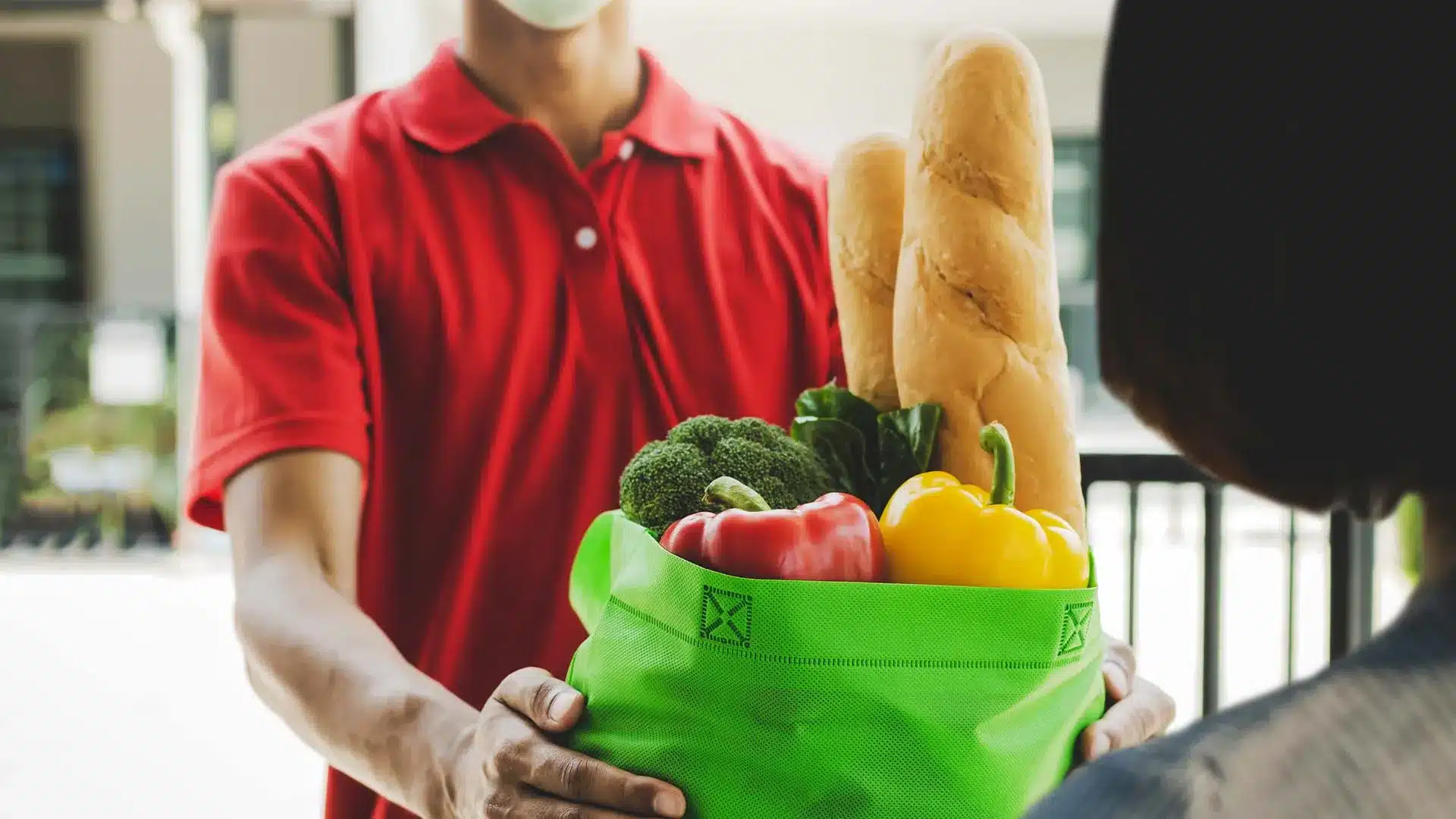 Photo of a man in a red t-shirt hand-delivering groceries in a green bag to a resident.